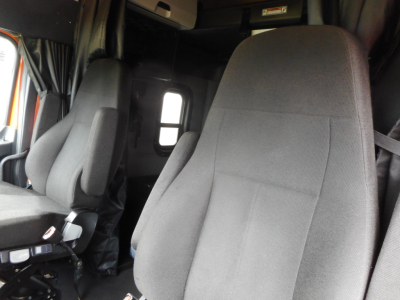 Cab interior of a Freightliner Cascadia showing driver and passenger seats and a small window; dark fabric seats.