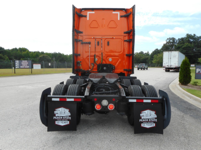 Rear view of a Freightliner Cascadia truck tractor with bright orange sleeper cab, dual rear axles, mud flaps with Peach State 50 Years logos, and view of fifth wheel and coupling area.