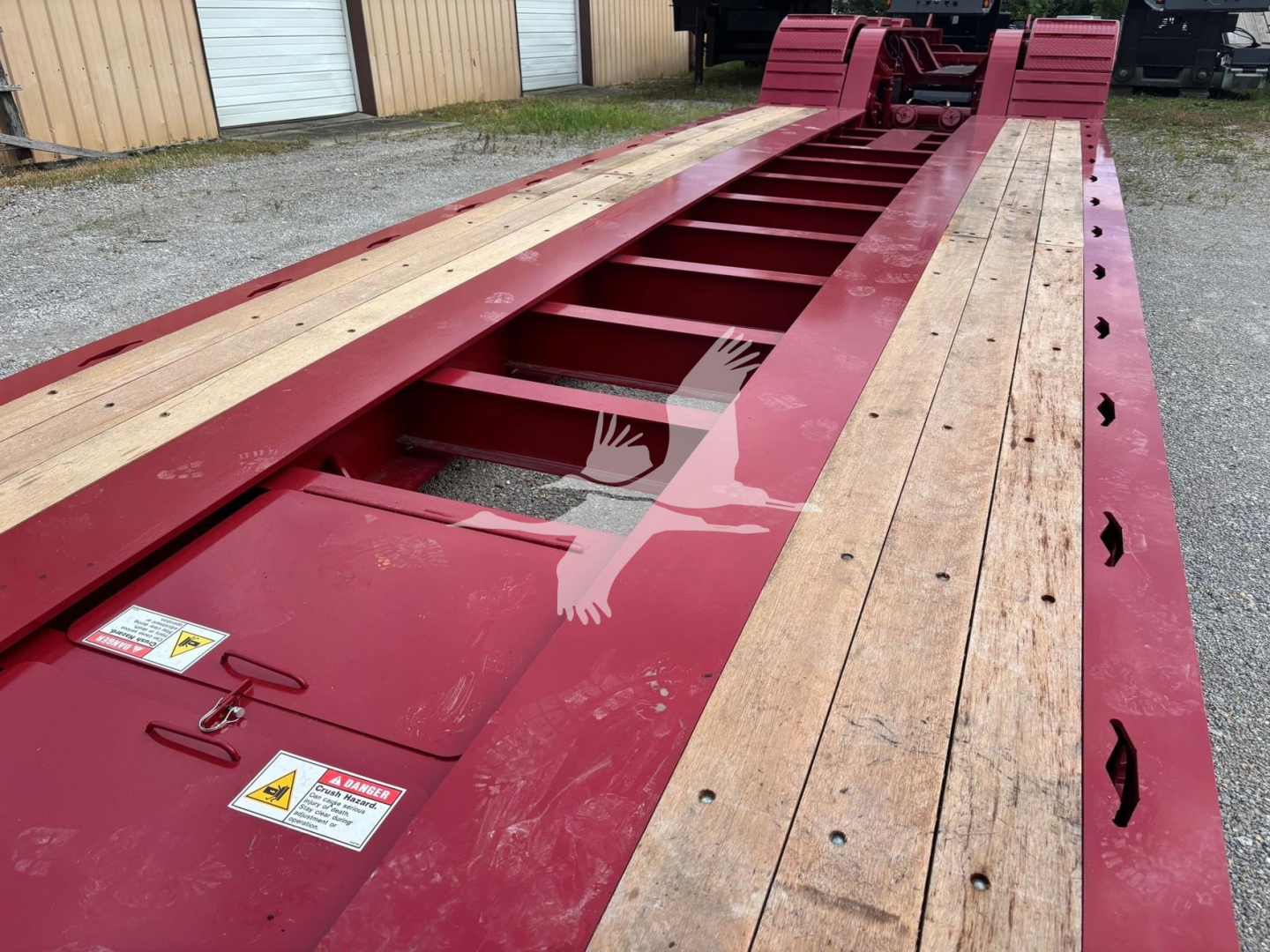 Lowboy trailer deck showing red steel frame with wooden planks on sides and center beam; warning labels and hinges near edge.