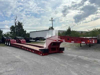 Trailer in a lot showing red Fontaine lowboy deck with gooseneck style front, dual rear axles, and heavy-duty landing gear; clear sky and industrial background.