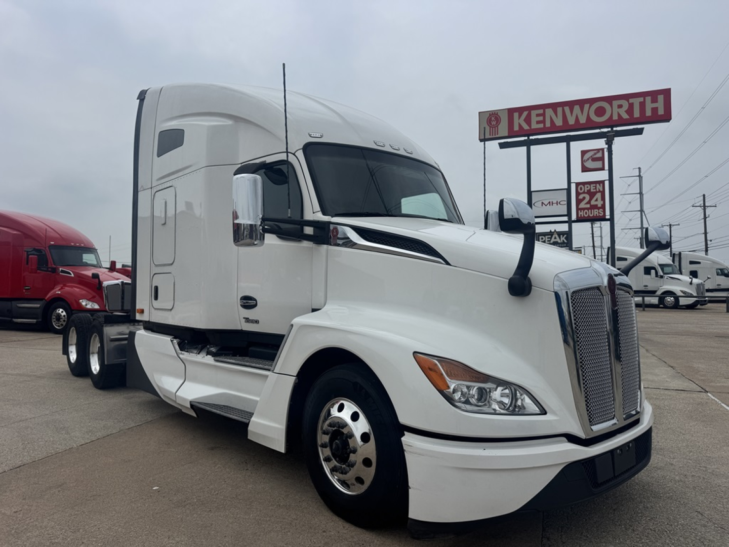 Kenworth T680 tractor-trailer in white with open Kenworth dealership sign in background. Visible text on image includes ‘KENWORTH’ sign, truck grille, side mirrors, and general outdoor lot.