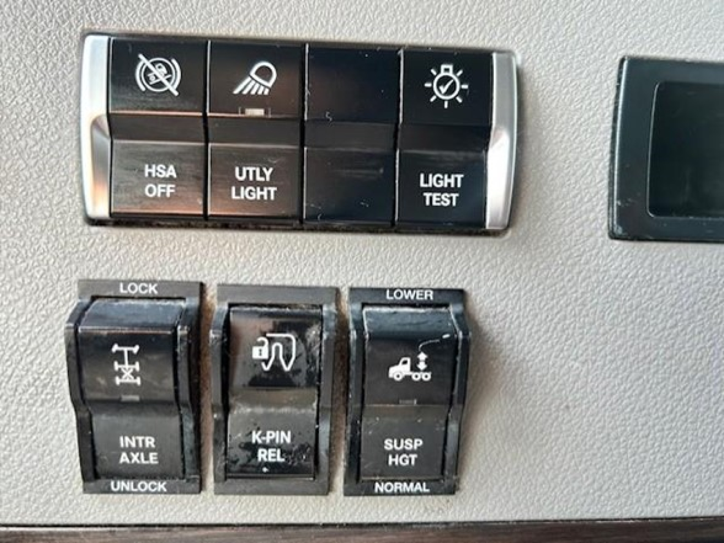 Dashboard switches and control buttons in a Freightliner Cascadia cabin showing various indicators and knobs with labels like HSA OFF, UTLY LIGHT, LIGHT TEST, LOCK, INTR AXLE UNLOCK, K-PIN REL, SUSP HGT, LOWER, and NORMAL.
