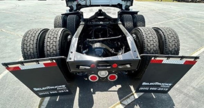 Trailer hitch of a Freightliner Cascadia 126 chassis showing dual rear axles and fifth wheel. Visible text on mud flaps reads 'SHELBY TRUCK LLC' and phone numbers, with reflective red/white tape.