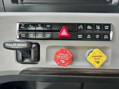 Dashboard of a Freightliner Cascadia showing HVAC/indicator buttons, red hazard triangle button, trailer brake lever, red trailer air supply knob, and yellow parking brake knob visible.