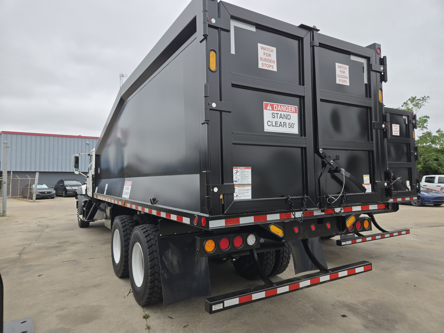 Rear view of a new 2026 Mack GR64B grapple truck showing dual rear tires, high-sided dump bed, back doors with warning labels, and reflective safety markings.