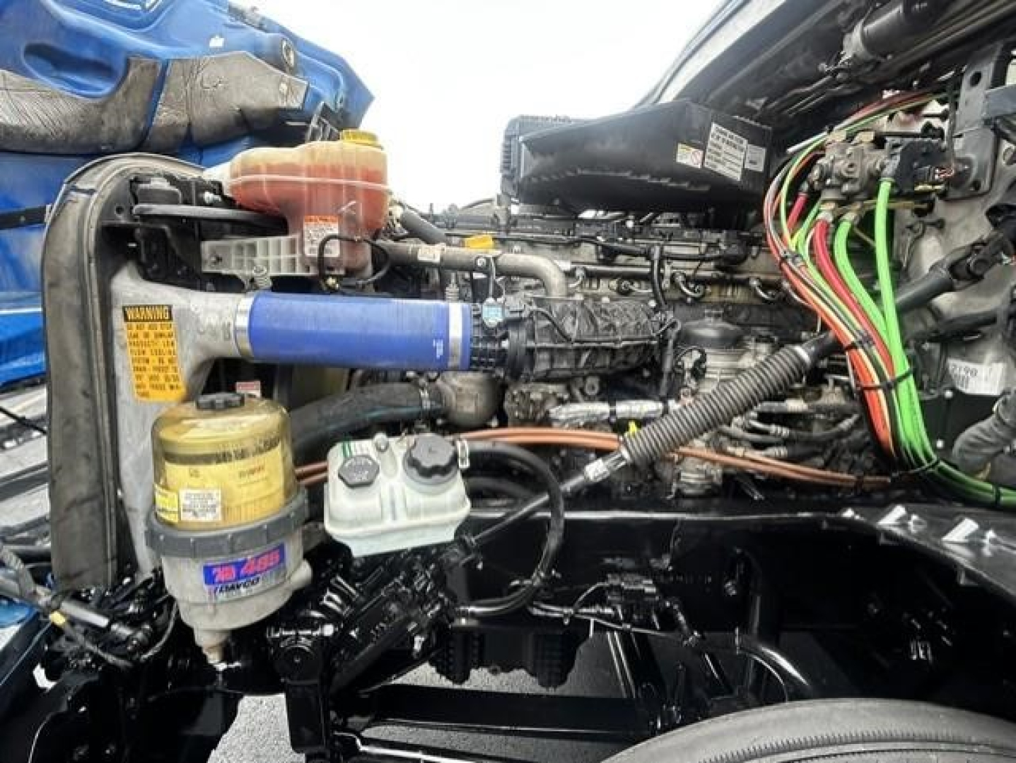 Diesel truck engine bay showing blue diagnostics hose, coolant reservoir, filter canister, and wiring harness in a Freightliner Cascadia engine bay.