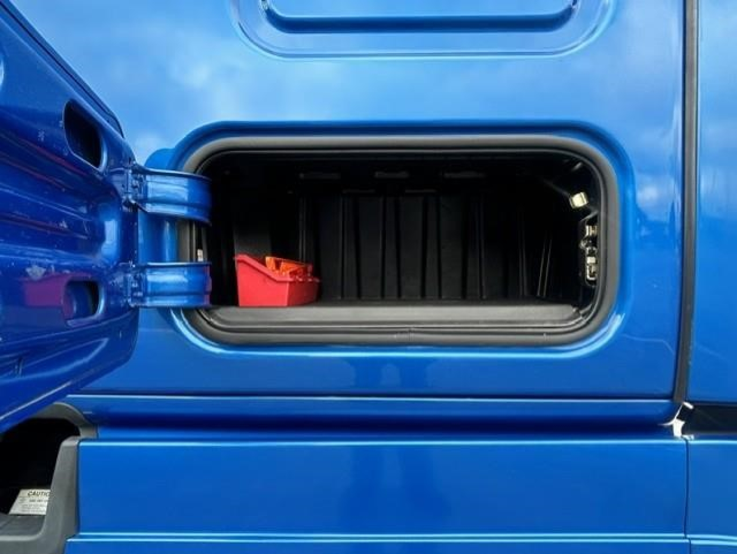 Truck side storage compartment open showing interior with a red item; blue Freightliner Cascadia 126 side panel