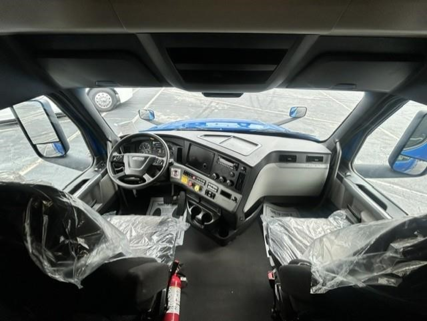 Cab interior of a Freightliner Cascadia left and right seats with plastic covers, dashboard, steering wheel, and controls visible.