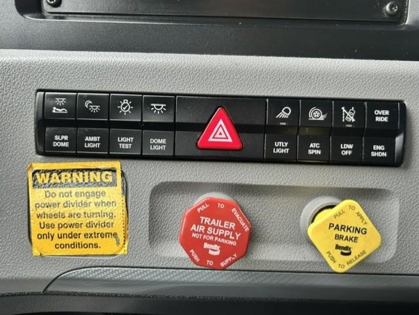 Opel Freightliner dash panel with hazard triangle switch, warning stickers, and control levers; visible labels include power/auxiliary lights, parking brake, trailer air supply, and parking brake yellow cap.