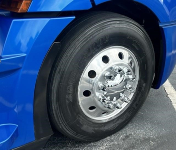 Front left wheel of a blue Freightliner Cascadia 126 sleeper truck showing tire, polished chrome hub, and fender.