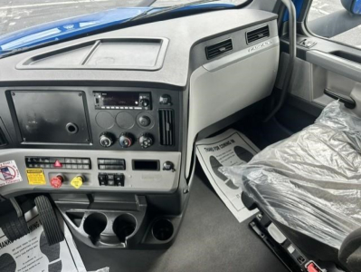 Cabin dashboard interior of a Freightliner Cascadia. Dashboard with radio, control knobs, vents, cup holders, and seat. Visible driver area and plastic seat cover.
