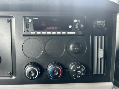 Truck dashboard panel showing FM radio display, climate controls, cigarette lighter, and vent; circular knobs and gauges visible in Freightliner Cascadia cabin.