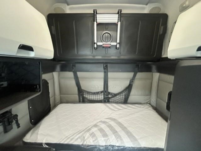 Freightliner Cascadia truck cab sleeper interior showing bunk bed with mattress, storage shelves, and netting; upper cabinet with latch and black panel.