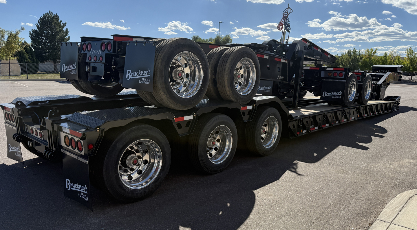 Lowboy trailer with dual-tandem axles and a heavy-duty gooseneck, three-axle spread, black finish, brand decals, and spare tires mounted on the frame; side view showing multiple axles and wheels.