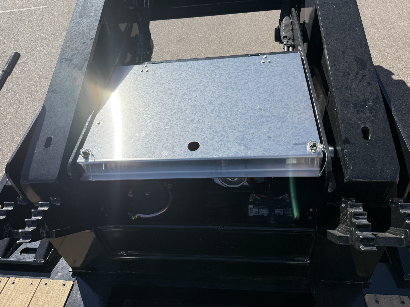 Equipment image of a trailer component showing a metallic rectangular access panel/box mounted in a black chassis with bolts, underside area and chain links visible.