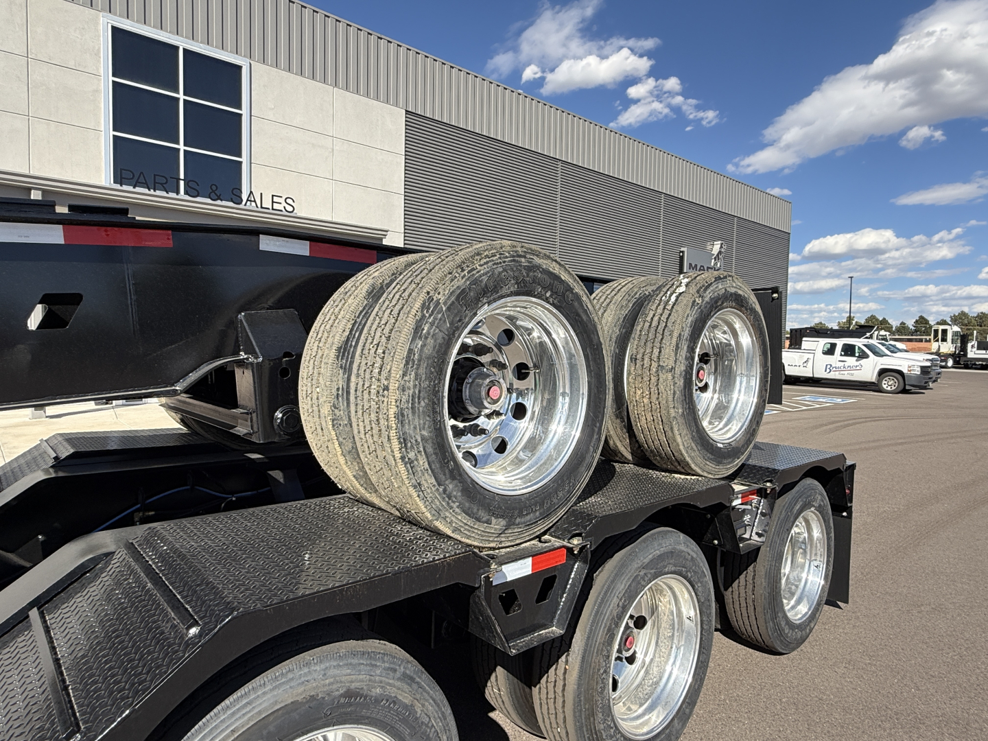 Trailer axles on a lowboy trailer showing dual tandem wheels and mud flaps, with chrome rims and treaded tires.