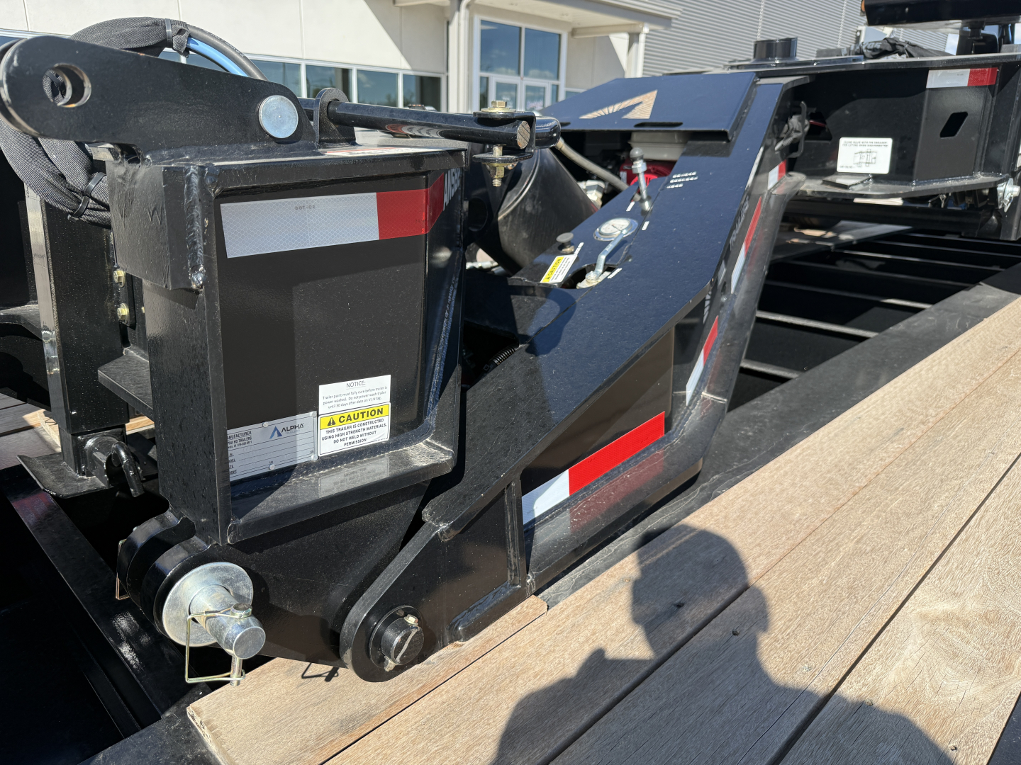 Trailer hitch and hydraulic pump assembly on a lowboy trailer, with safety labels, mechanical arms, and wooden deck planks visible.