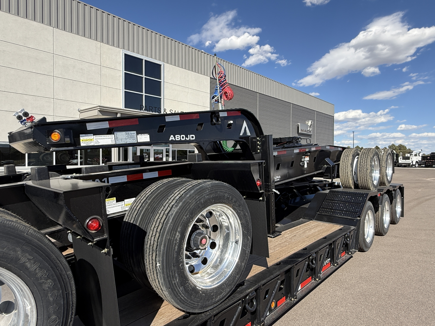 Trailer showing a multi-axle lowboy deck with spare wheels and tires, black frame, reflective tape, and a red safety chain; visible label and components along the trailer