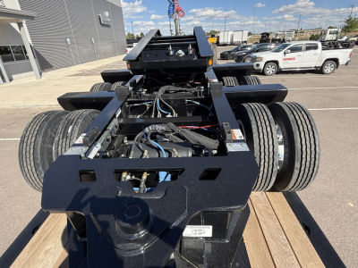 Alphanumeric trailer chassis showing hydraulic and electrical components, dual rear axle with tires, black powder-coated frame, and mounting plate with wiring harnesses.
