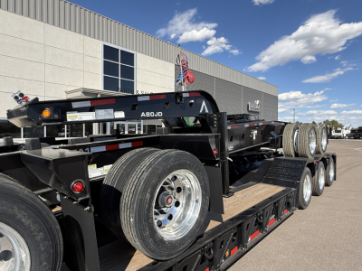 Trailer showing a multi-axle lowboy deck with spare wheels and tires, black frame, reflective tape, and a red safety chain; visible label and components along the trailer