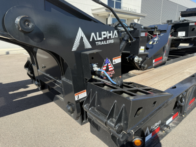 Alpha Trailers logo on black heavy-duty trailer with metal frame and hydraulic components; close-up showing part of the frame and platform.