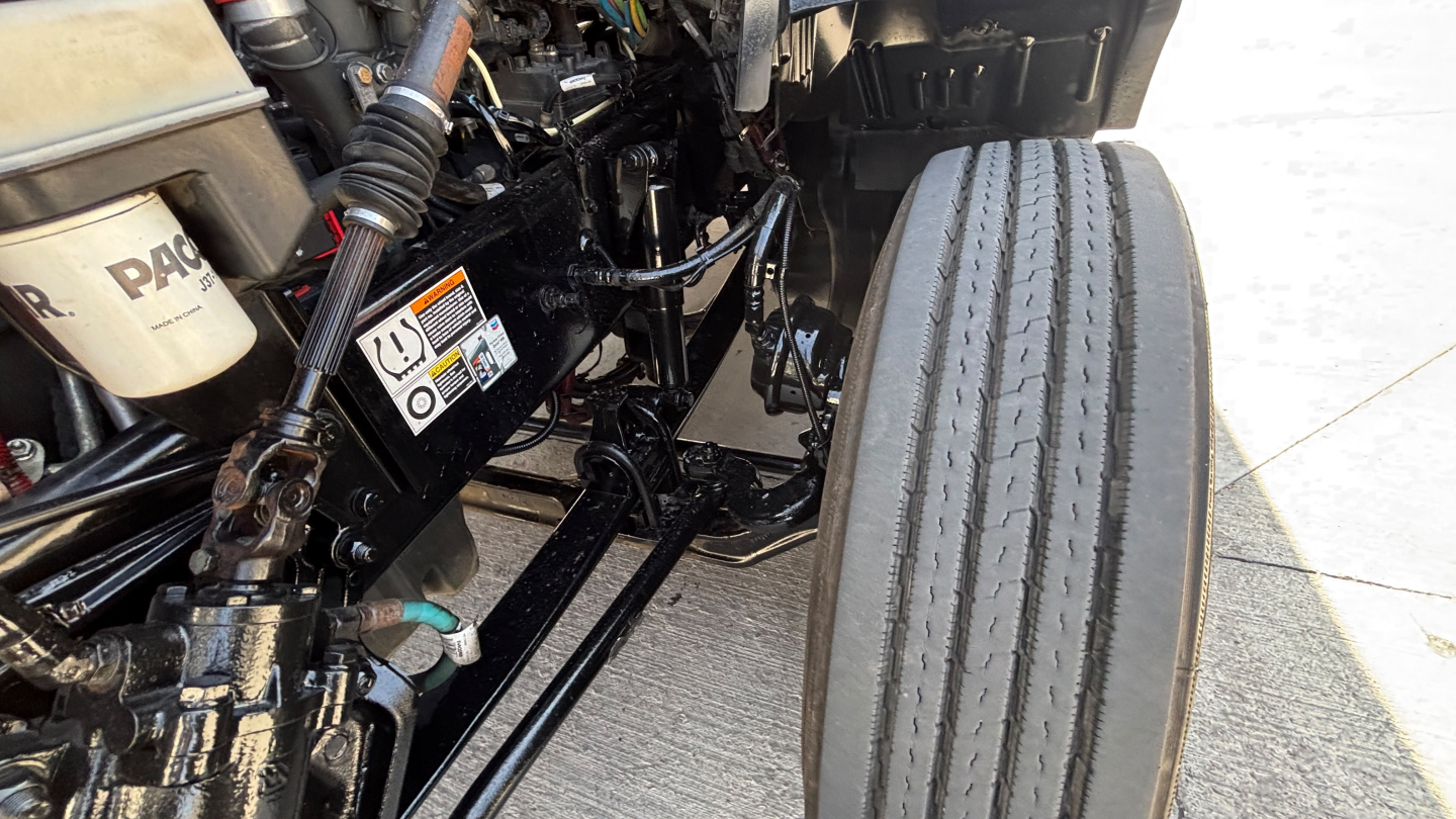 Truck engine bay area with front axle, suspension, and header pipes visible; front tire and chassis components in view.