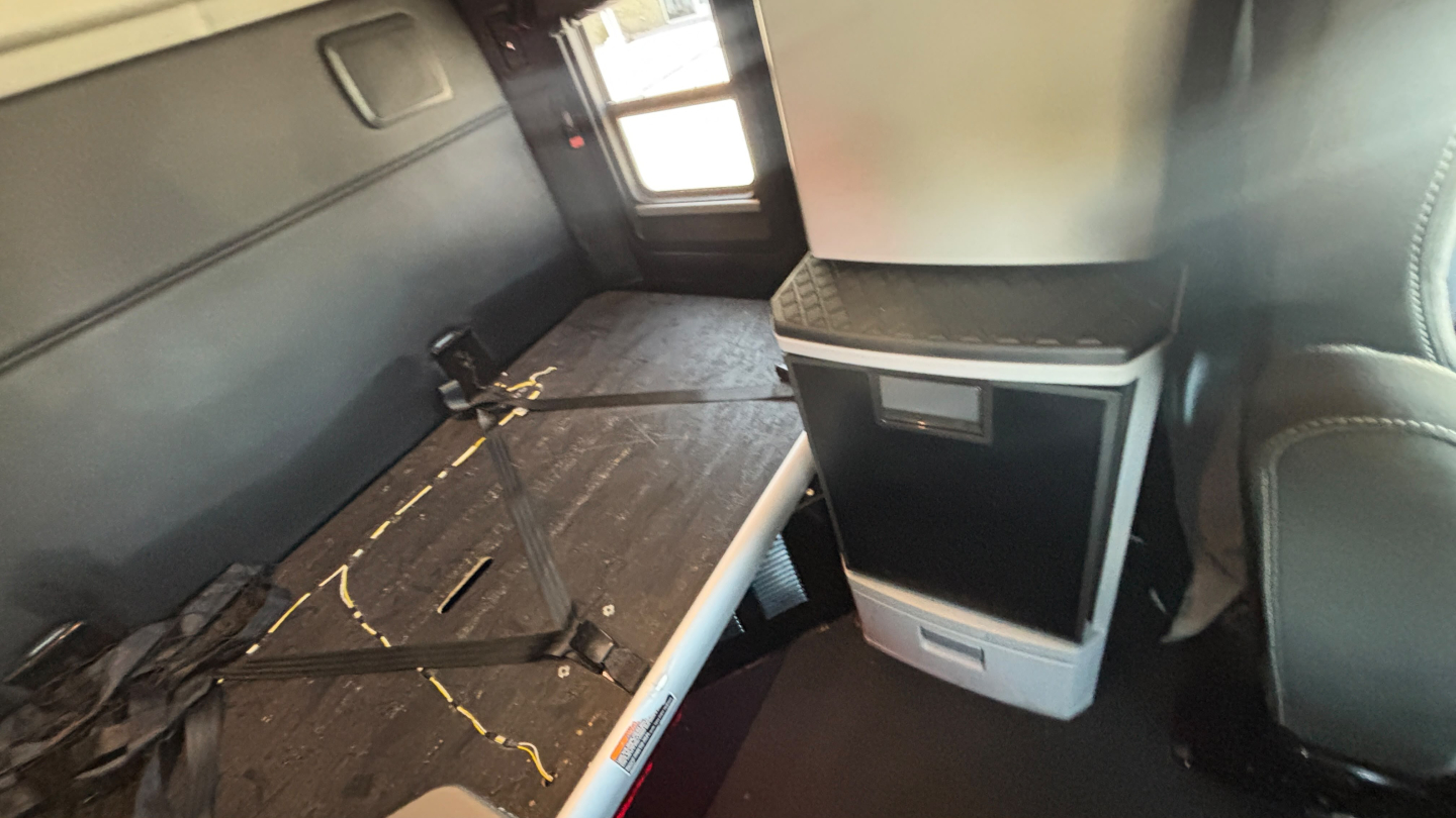 Kenworth T680 truck interior view showing sleeper cab area with seat, seating belt, and storage cabinet; cabin floor shows scattered debris with seat belt buckle and door outline visible.