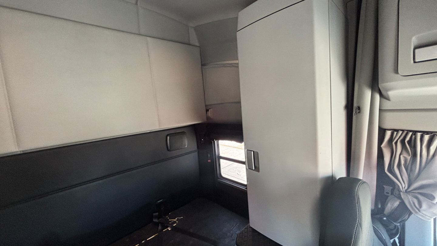Cab interior of a 2023 Kenworth T680 sleeper, showing a compact berth with light-colored upholstery, window, and storage compartments.