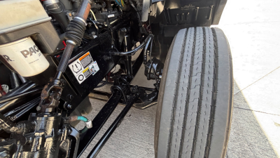 Truck engine bay area with front axle, suspension, and header pipes visible; front tire and chassis components in view.