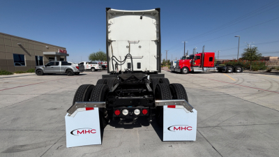Tractor truck rear view showing fifth-wheel connection and dual rear axles with white MHC mud flaps, view from a parking lot.