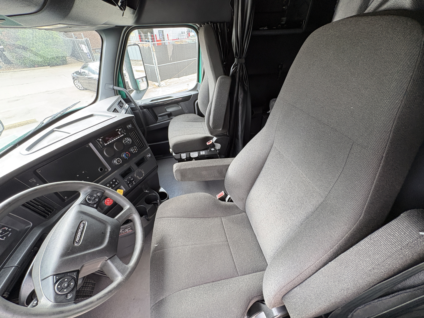 Truck cab interior showing driver's seat and dashboard of a Freightliner Cascadia 126 with steering wheel and controls visible.
