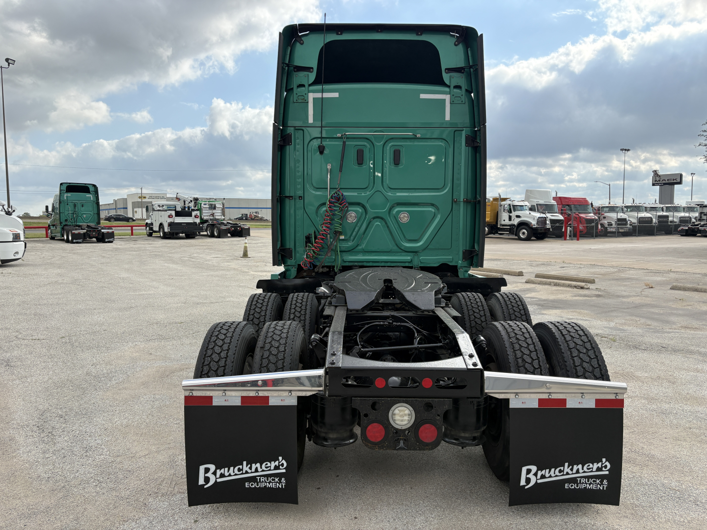 Tractor truck rear view showing fifth-wheel coupling on a Freightliner Cascadia 126, dual rear axles, green sleeper cab, mud flaps with Bruckner’s logo, parking lot.