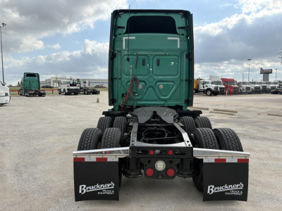 Tractor truck rear view showing fifth-wheel coupling on a Freightliner Cascadia 126, dual rear axles, green sleeper cab, mud flaps with Bruckner’s logo, parking lot.