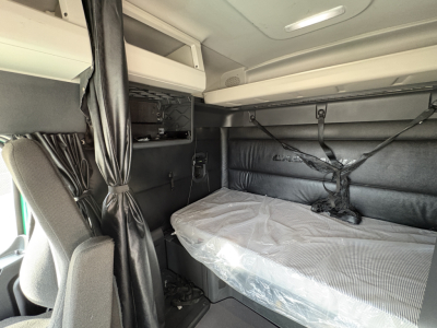Cab interior of a Freightliner Cascadia 126 sleeper showing bunk bed with mattress, overhead storage, and straps.