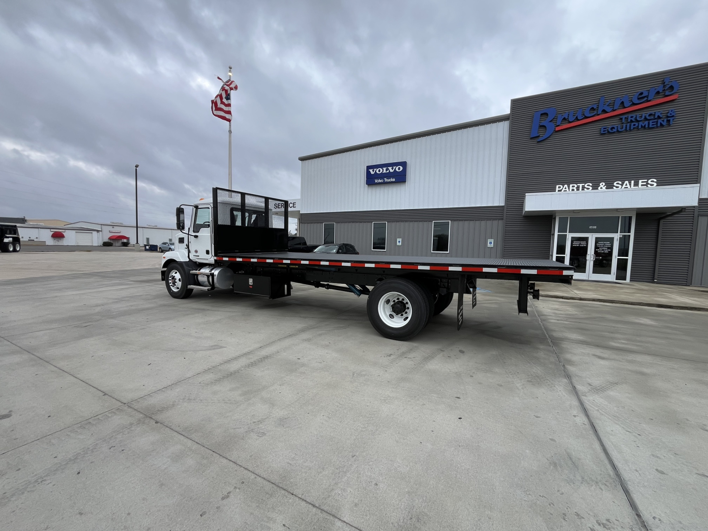 Flatbed truck in front of dealer building showing 2026 Mack MD6 with long flatbed and black-and-red reflective safety stripes, white cab, dealership signage in background.