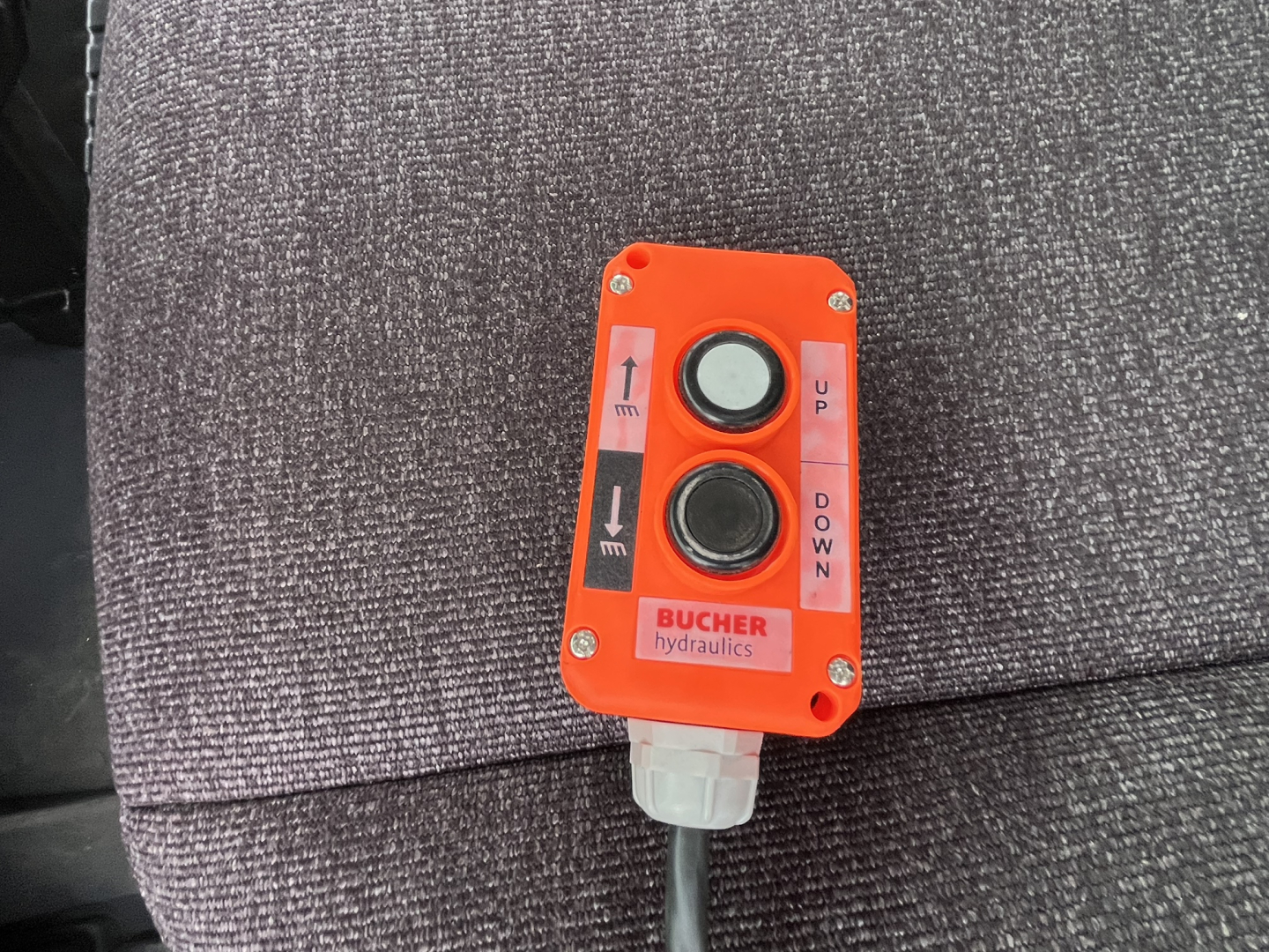 Orange Bucher hydraulic control box with UP/DOWN arrows on lever and hydraulic hoses, mounted on gray fabric truck seat.