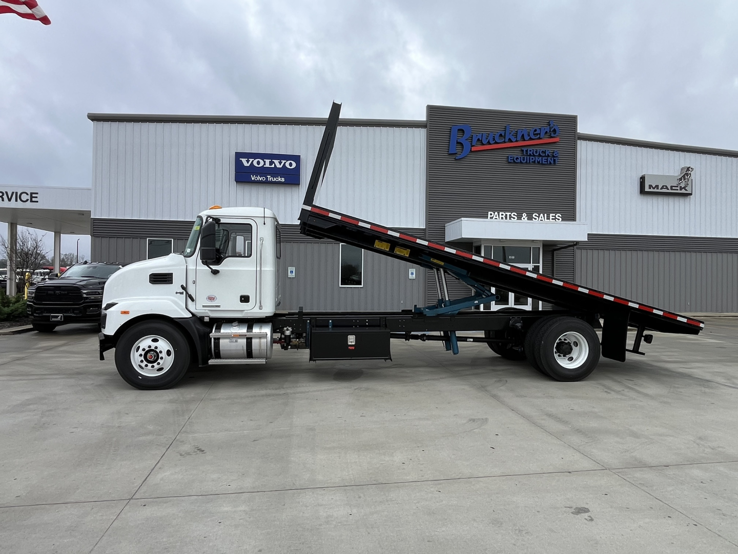 Flatbed tow truck in front of a service shop with Mack MD6 logging on the sign and a long tilt flatbed.