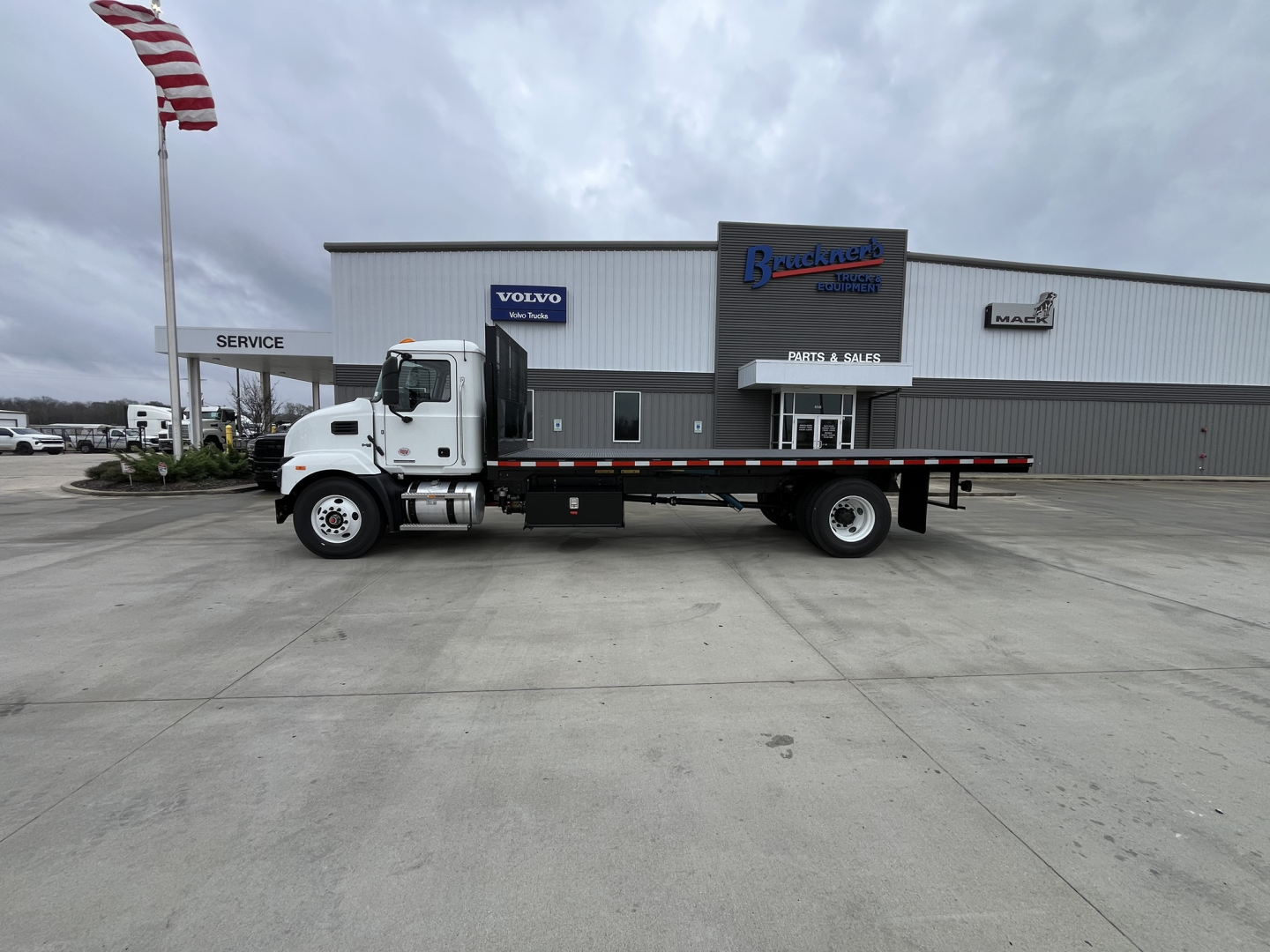 Flatbed truck in dealership lot with Mack MD6 cab and long flatbed, white cab, gray/dark flatbed, building in background with brand logos.