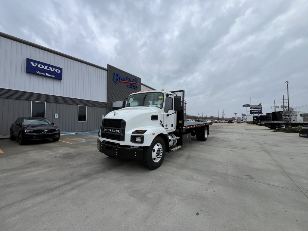 2026 Mack MD6 flatbed truck front-left view with white cab and flatbed, dealership building in background showing Volvo sign and other branding.