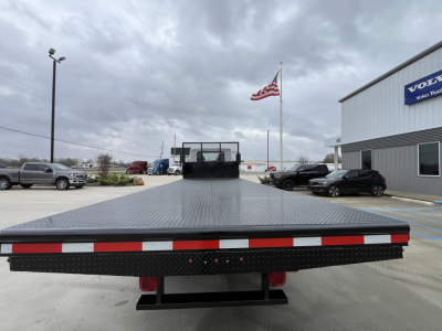 Flatbed truck bed of a 2026 Mack MD6 with diamond plate surface, rear view showing taillights and metal ramp. Visible text on sticker area: none legible.