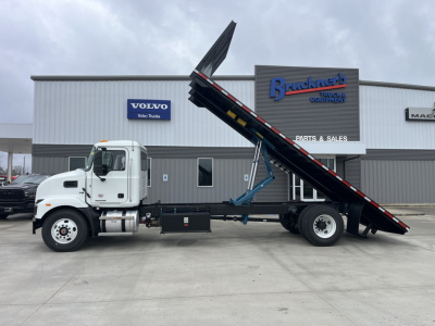 Truck body with a raised tilting flatbed on a Mack MD6 chassis, in a dealership lot with Brackett Truck Equipment signage and Volvo logo in background.