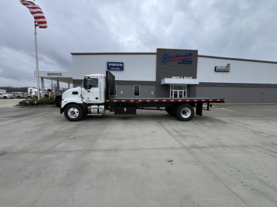 Flatbed truck in dealership lot with Mack MD6 cab and long flatbed, white cab, gray/dark flatbed, building in background with brand logos.