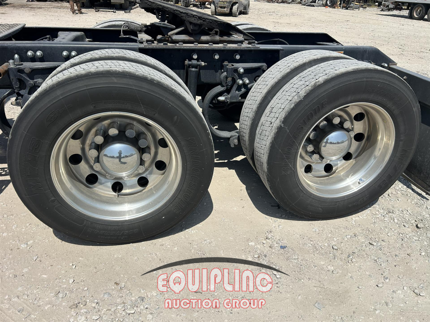 Kenworth W900 wheel pair showing dual rear axles with polished steel rims and hubcaps, undercarriage metal frame, gravel surface.