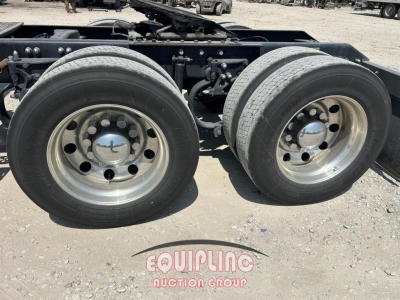 Kenworth W900 wheel pair showing dual rear axles with polished steel rims and hubcaps, undercarriage metal frame, gravel surface.