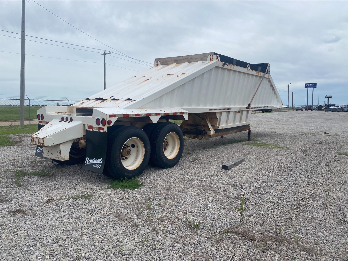 Dump trailer with raised bed and dual rear wheels, worn white paint, rust spots, and a sloped-sided steel dump body in a gravel lot.