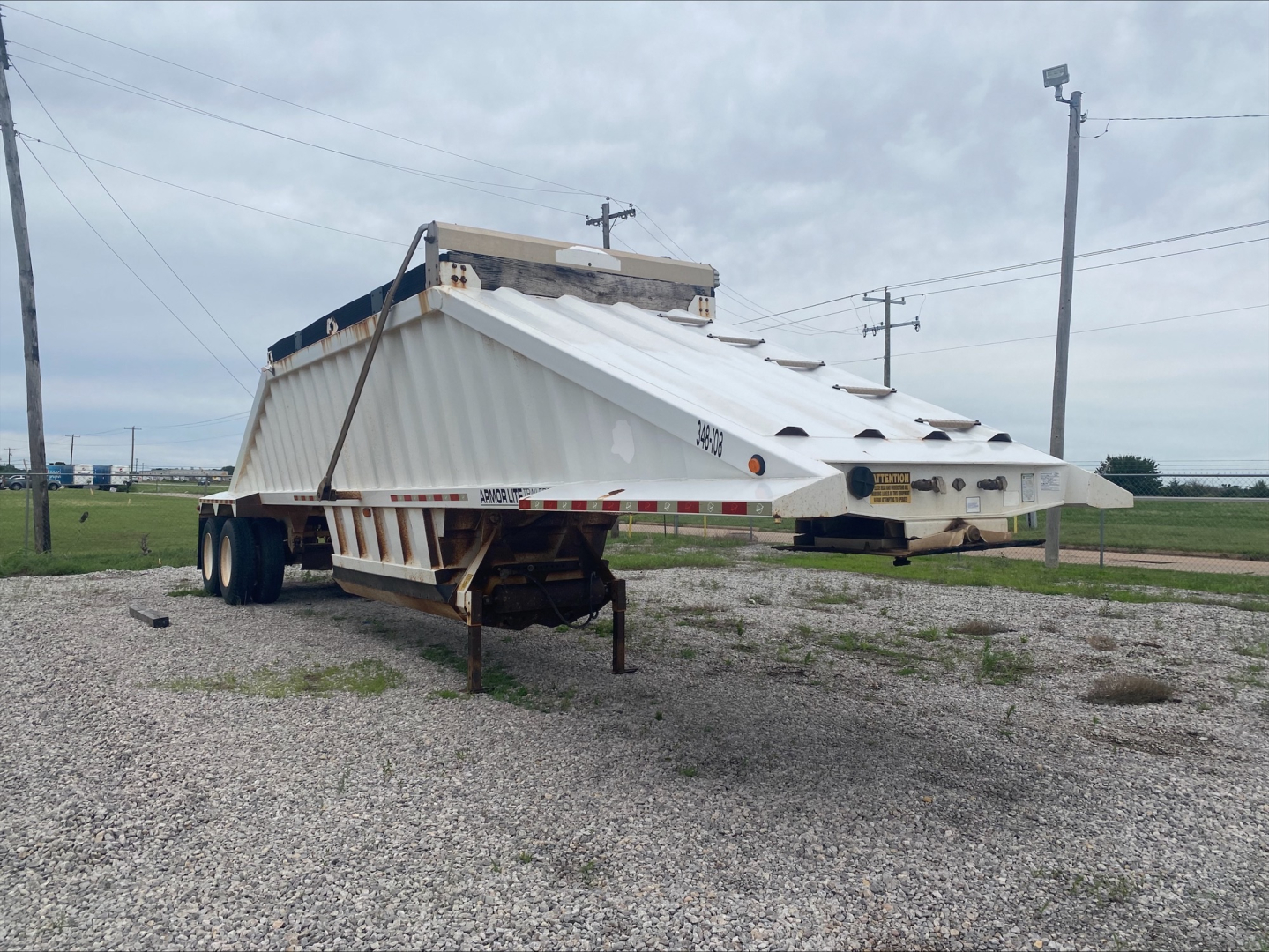 Dump trailer in a gravel lot showing a raised armored body with white sides and a skinned triangular beavertail.