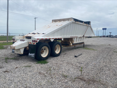 Dump trailer with raised bed and dual rear wheels, worn white paint, rust spots, and a sloped-sided steel dump body in a gravel lot.