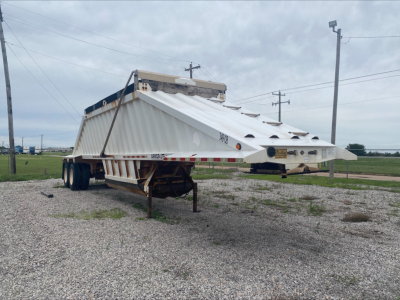 Dump trailer in a gravel lot showing a raised armored body with white sides and a skinned triangular beavertail.