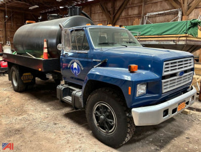 Ford F-800 septic tank vac truck in blue with a large black cylindrical tank on a flatbed, orange warning lights, logo on door, inside a wooden barn.