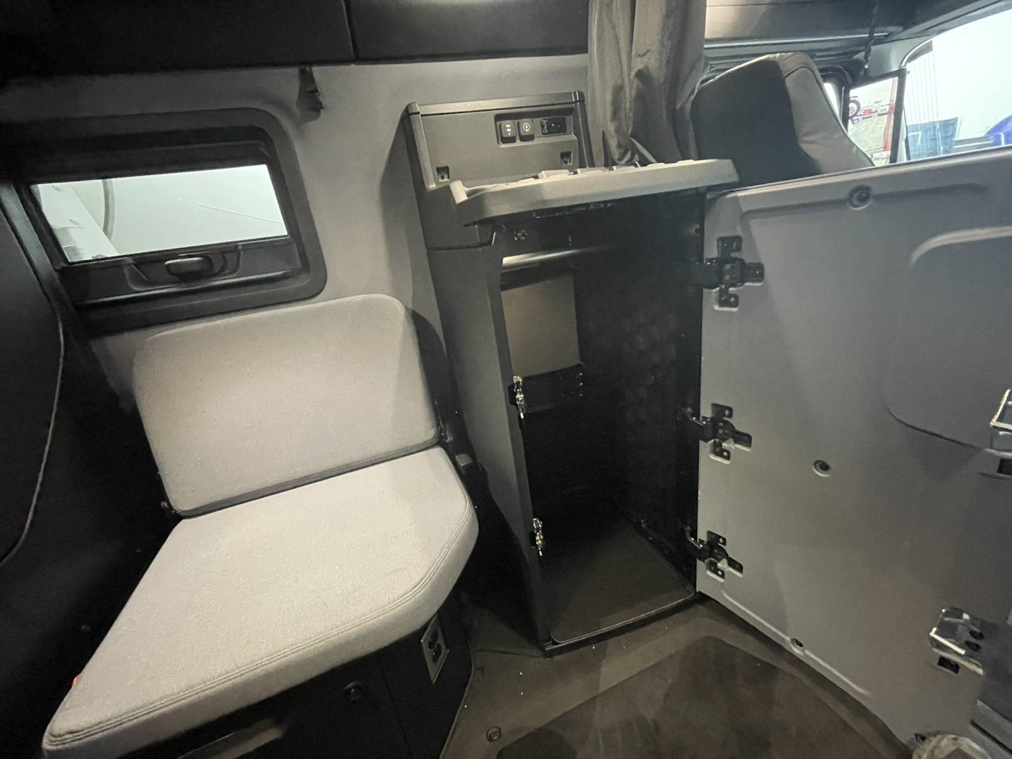 Truck cab interior showing a small black cabinet with shelves and an open door, gray seat, and parts of the sleeper area.